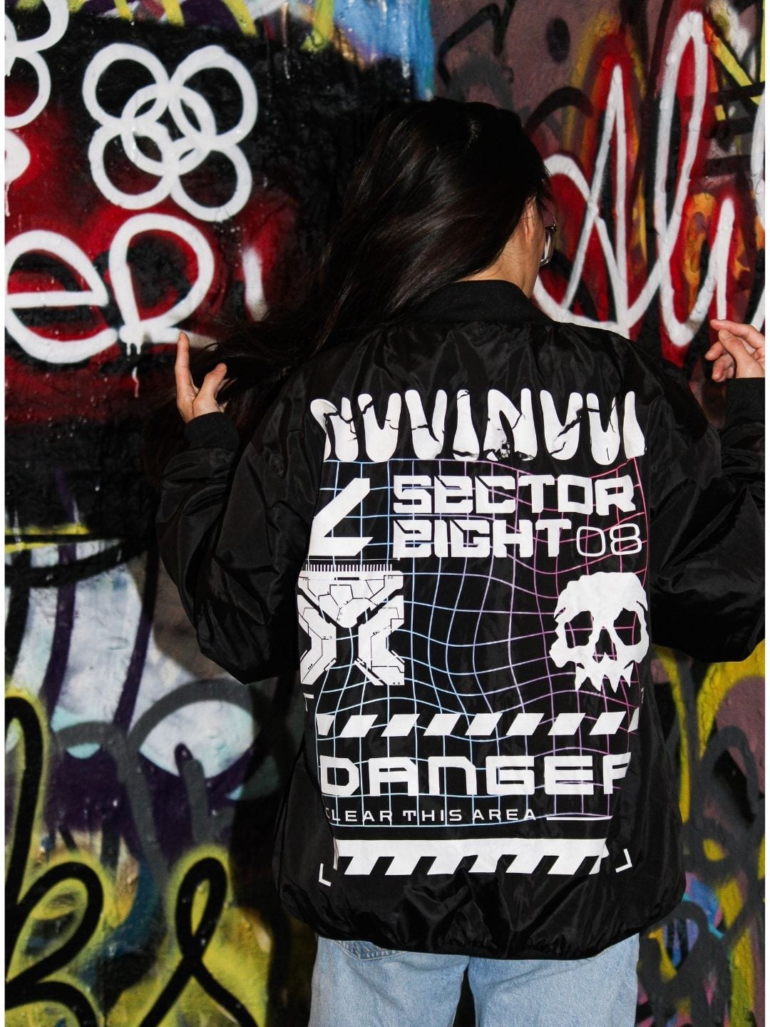 Bomber noir porté de dos, impression dorsale blanche avec crâne, grille filaire et bandeau DANGER, devant un mur de graffitis.